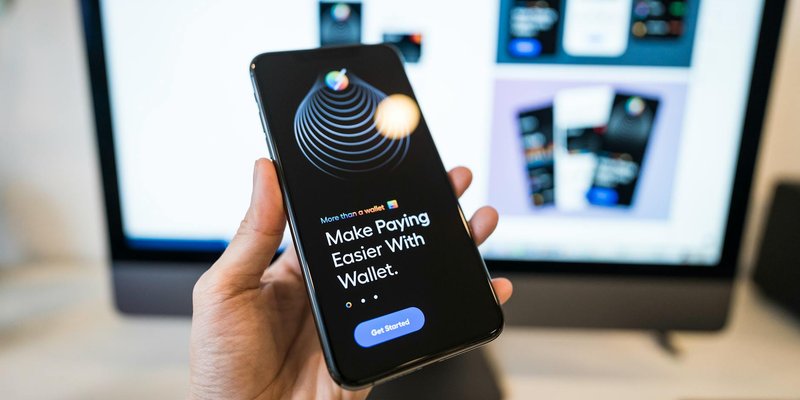 Hand holding smartphone displaying digital wallet app interface, blurred monitor in background.