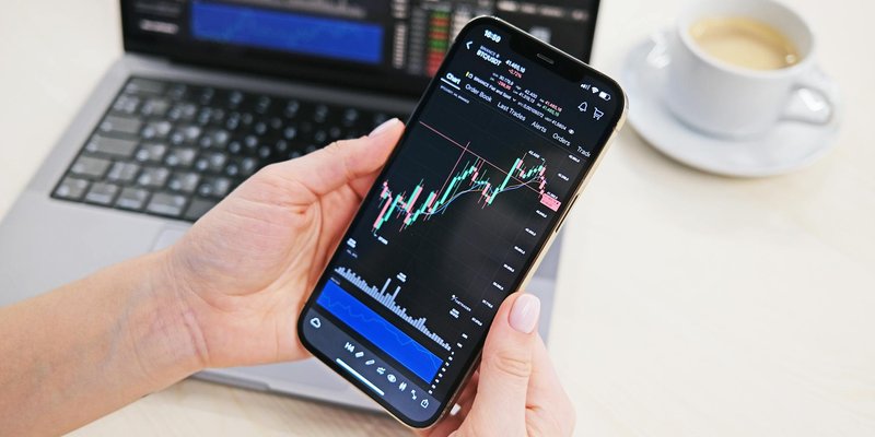 Close-up of hands holding smartphone displaying stock charts with laptop in the background on a desk