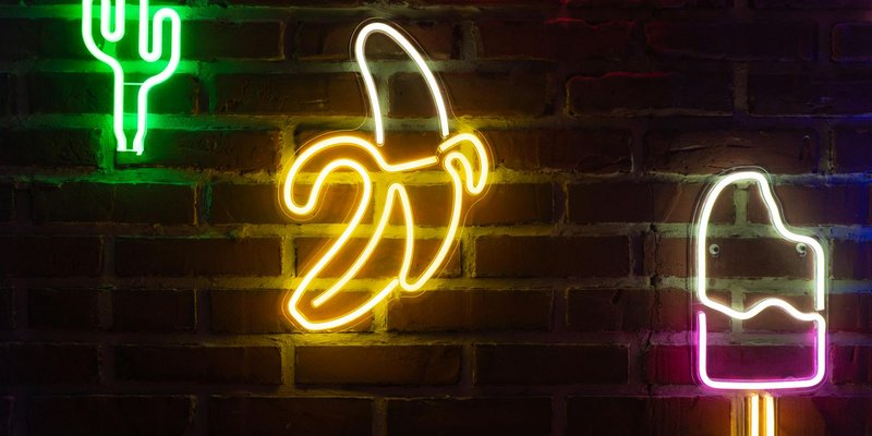 Colorful neon signs of banana, cactus, and ice cream pop against a brick wall.