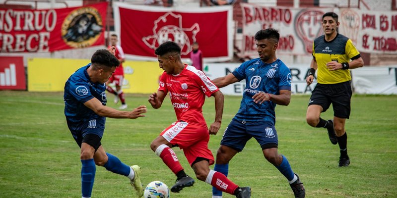 Soccer players in action during a competitive match on a grass field with spectators in the stands.
