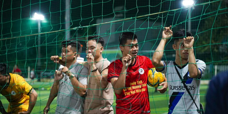 Asian men standing behind netting on a soccer field at night, holding a soccer ball.