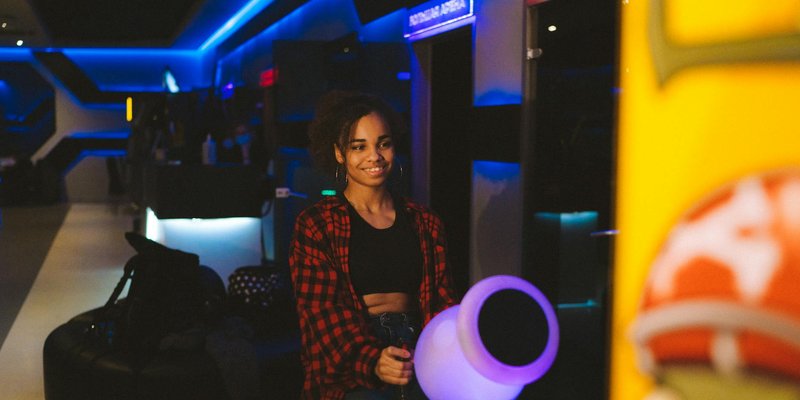 A woman enjoys playing an arcade game in a neon-lit setting, smiling at the gaming machine.